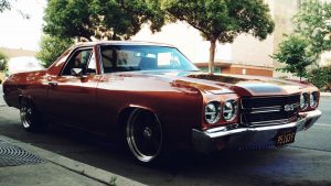 Red Chevrolet El Camino parked on a tree-lined city street, evoking a nostalgic and retro feel.